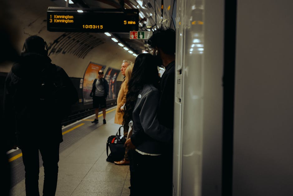 A group of commuters standing on the platform of Kennington Station, with some wearing dark clothing and carrying backpacks or bags, waiting for the train. The station's curved ceiling and illuminated