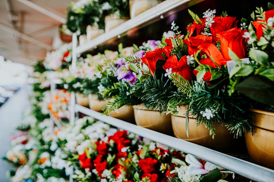 A row of beige flower pots filled with vibrant floral arrangements sits on a metal shelf at a flower stall, featuring bright red roses, white flowers, and assorted green foliage. The flowers are fresh