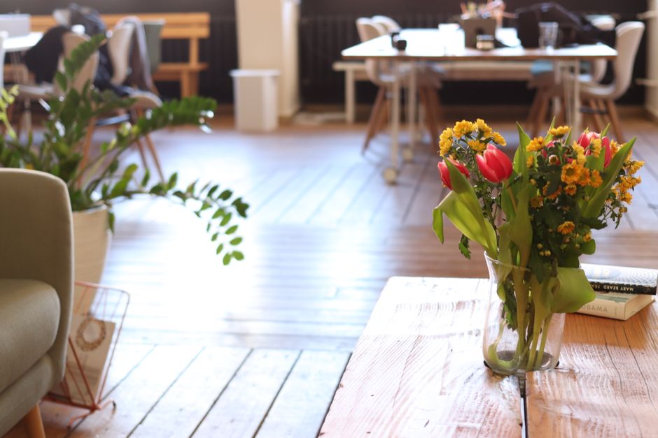 A clear glass vase filled with a vibrant floral arrangement featuring red tulips, small yellow chrysanthemums, and green leaves sits on a light wooden table inside a modern, well-lit café. The arrang