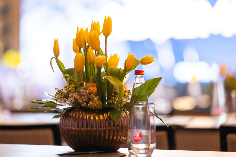 A floral arrangement featuring a bouquet of vibrant yellow tulips with closed and partially open petals, arranged in a round, dark amber glass vase with vertical ridges. The bouquet is complemented by