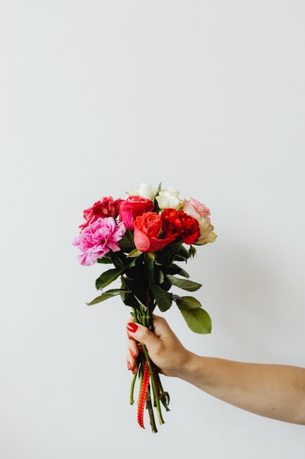 A hand holding a fresh bouquet of mixed flowers including pink, red, and white roses, along with pink carnations, arranged in a natural style with green foliage. The bouquet is bound with a red ribbon