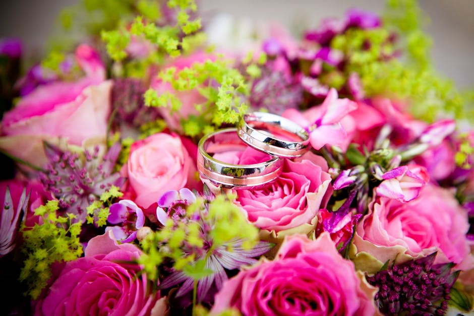 A close-up view of a floral arrangement featuring pink roses, light green hydrangeas, and purple accent flowers, with two polished silver wedding rings resting on the bouquet. The flowers appear fresh