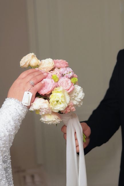 A close-up of a bridal bouquet being held by a bride in a white lace dress, with her hand displaying a diamond ring and a wedding tag. The bouquet consists of fresh pink and cream roses, lisianthus wi