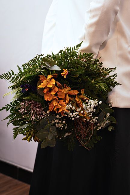 A person dressed in a white jacket and black skirt holds a large, lush floral bouquet featuring a mix of fresh flowers and greenery. The arrangement includes vibrant orange blossoms, delicate white sp