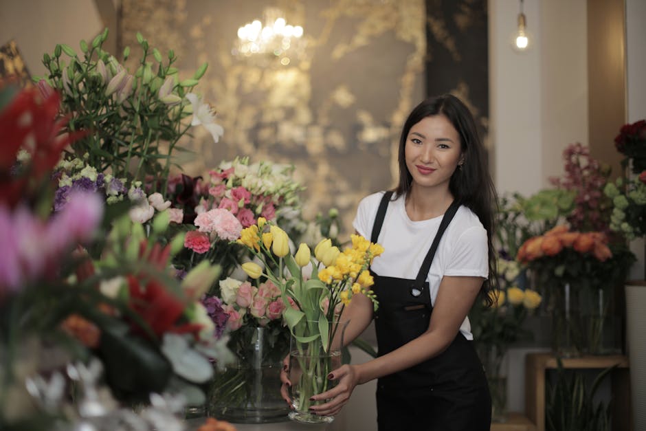 A young female florist with long dark hair, wearing a white t-shirt and black apron, standing in a floral shop surrounded by an assortment of colorful fresh flowers, including yellow tulips, pink rose