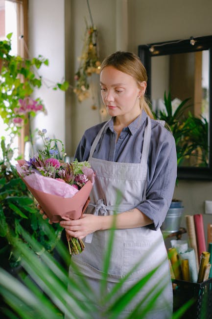 A woman in a blue shirt and light grey apron stands indoors holding a bouquet of fresh flowers, including pink and purple blooms wrapped in pink paper. She appears to be examining the floral arrangeme