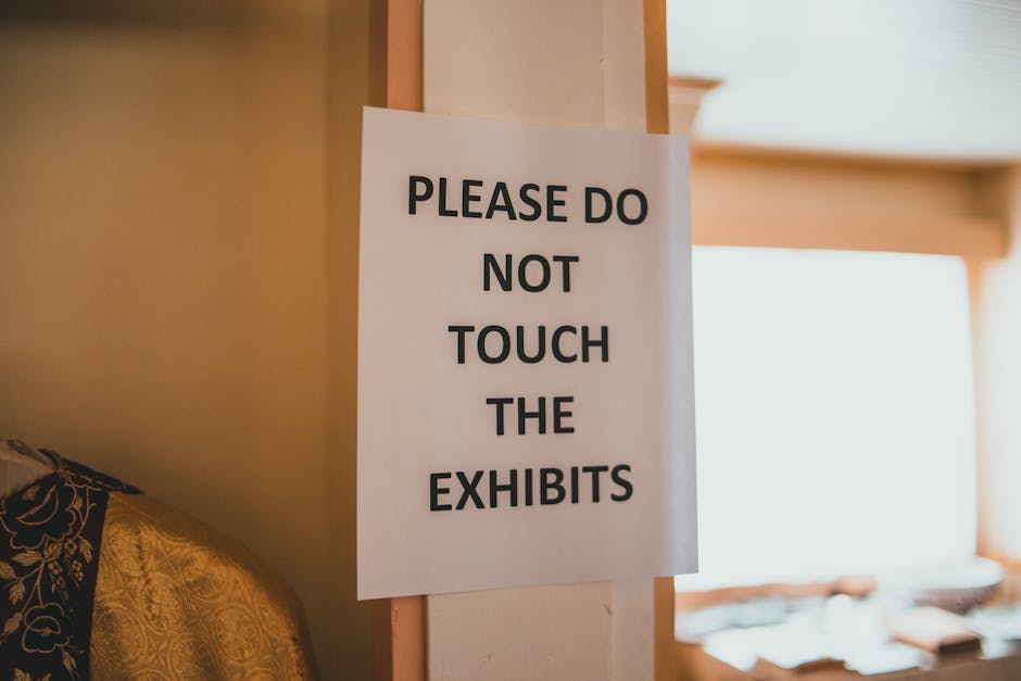 A close-up of a white paper sign with bold black text reading 'PLEASE DO NOT TOUCH THE EXHIBITS' affixed to a beige wall inside a room. Part of a patterned fabric upholstery is visible on the left sid