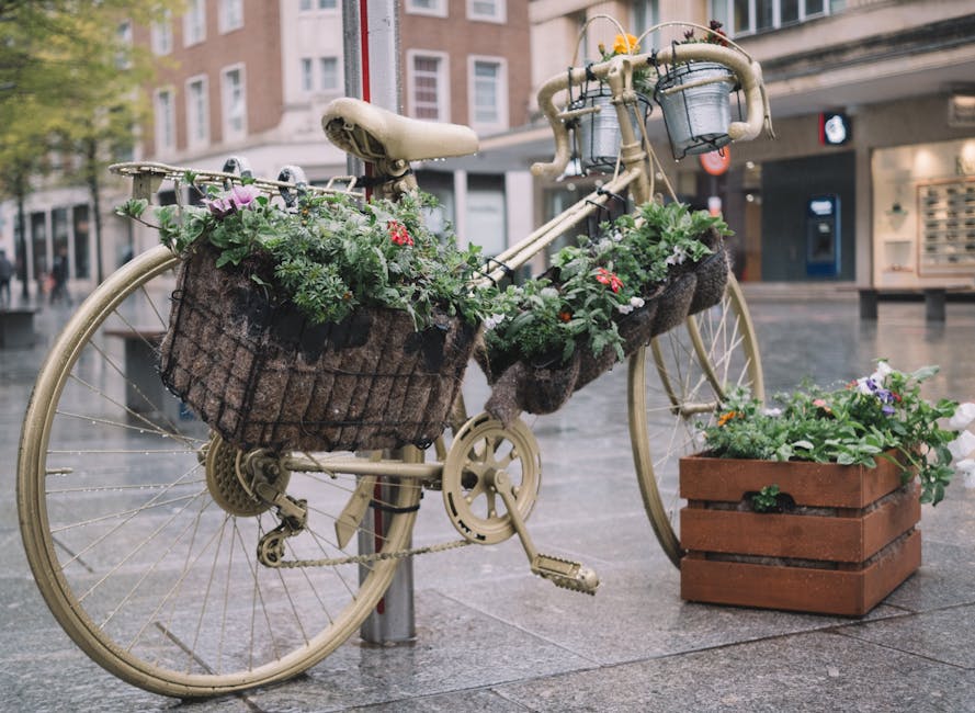 A vintage-style beige bicycle with a large wire basket mounted on the front, filled with an arrangement of fresh flowers including pink roses, red and purple blooms, and lush green foliage. The bicycl