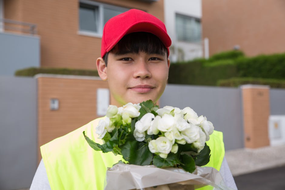 A young male delivery person wearing a red cap and a high-visibility yellow vest is smiling and holding a bouquet of fresh white flowers, possibly white roses or spray roses, wrapped in white paper. T