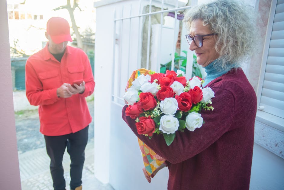 A woman with curly gray hair, wearing glasses, a light blue turtleneck, and a maroon sweater, holds a bouquet of fresh red and white roses wrapped in colorful paper. She stands near a white window shu